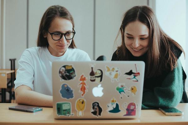 Two colleagues at a desk, looking together at the screen of a sticker-covered laptop.